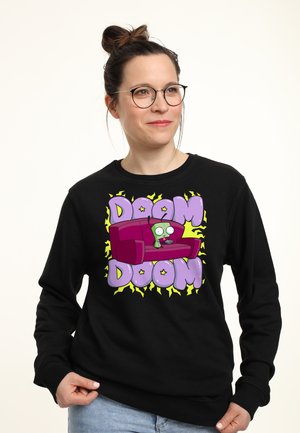 Vrouw met bril en zwart sweatshirt met cartoon alien op paarse bank en tekst "DOOM DOOM" op paarse en gele achtergrond.