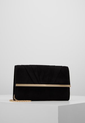 Bolso de mano negro de ante con detalle de barra metálica dorada y correa de cadena dorada, colocado sobre una superficie blanca con un fondo gris.