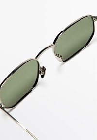 Ovale solbriller med grønne linser og en slank sølvmetalramme. Fremhævet af markante næsepuder og tynde stænger.