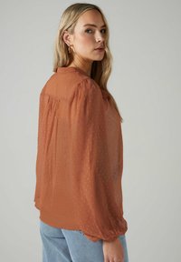 Burnt orange, sheer blouse with a dotted texture, gathered shoulder seams, and long, puffed sleeves. Worn with light blue jeans.