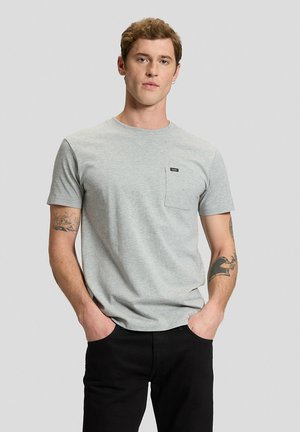 Uomo con capelli corti e tatuaggi indossa una t-shirt grigia chiara con taschino sul petto e pantaloni neri, in piedi con le mani in tasca su uno sfondo neutro.