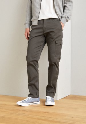 Veste bomber grise matelassée sur une chemise blanche, associée à un pantalon cargo vert olive avec des poches latérales et complétée par des baskets en toile bleues.