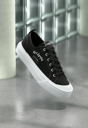 Zapatilla deportiva negra de Karl Lagerfeld de perfil bajo con suela y cordones blancos, con texto de la marca y puntera de goma, exhibida sobre suelo gris reflectante.