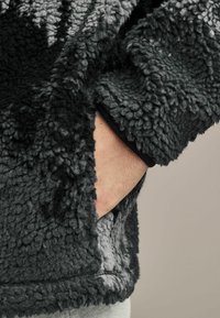 Polarowy kurtka w gradientowych odcieniach czerni i szarości, z teksturowaną powierzchnią i boczną kieszenią z gładkim wykończeniem.