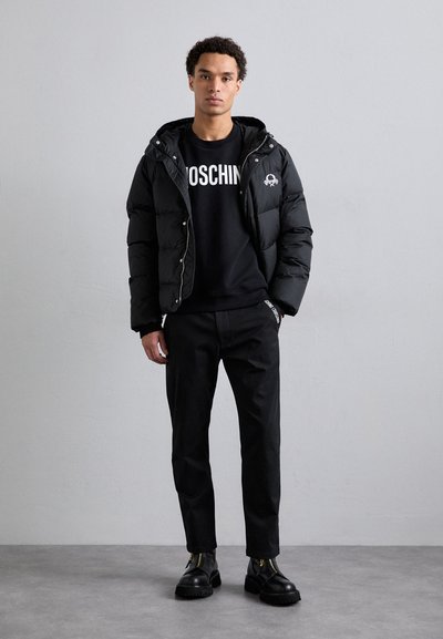 Schwarze Pufferjacke mit Kapuze und Reißverschluss, getragen über einem schwarzen Sweatshirt mit weißem Text. Schwarze Hose und schwarze Stiefel mit Reißverschluss runden das Outfit ab.