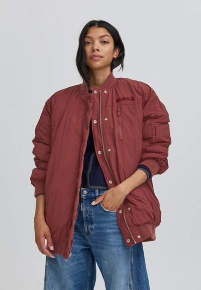 Bordo krāsas pārklātais bomber jakas ar rāvējslēdzēja krūšu kabatu, pogu aizvēršanas sistēmu, ribotu aproču un gludu tekstūru. Vilkta virs zila augšdaļas un džinsiem.