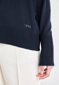 Granatowy sweter z prążkowanymi mankietami i dołem, z małym logo "BOSS", noszony na białych, fakturowanych spodniach.