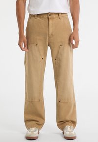 Beige Cargo-Hosen aus Baumwolle, mit weiten Beinen, vorderen Taschen mit Klappen und metallischen Druckknopf-Details. Kombiniert mit weißen Sneakern.