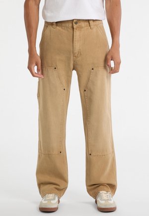 Beige cargo pants made of cotton, featuring wide legs, front pockets with flaps, and metal snap accents. Paired with white sneakers.