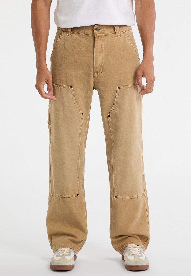 Beige Cargo-Hosen aus Baumwolle, mit weiten Beinen, vorderen Taschen mit Klappen und metallischen Druckknopf-Details. Kombiniert mit weißen Sneakern.