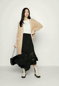 Cardigan long beige avec des finitions blanches, haut blanc, jupe noire à volants superposés, et bottines crème. Le mannequin tient un sac à main noir.