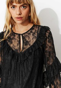 Blouse en dentelle noire avec col en V orné de volants, manches longues à volants superposés, et fermeture par bouton à l'encolure avec une fente clé.