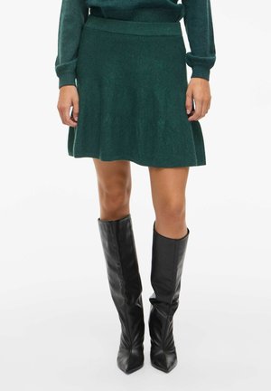 Femme portant une jupe en tricot vert foncé et un pull assorti, associés à des bottes en cuir noires montant jusqu'aux genoux, se tenant devant un fond blanc.