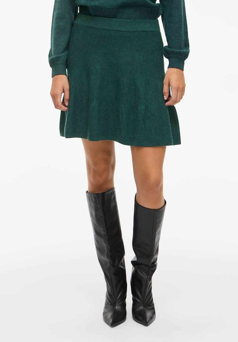 Femme portant une jupe en tricot vert foncé et un pull assorti, associés à des bottes en cuir noires montant jusqu'aux genoux, se tenant devant un fond blanc.