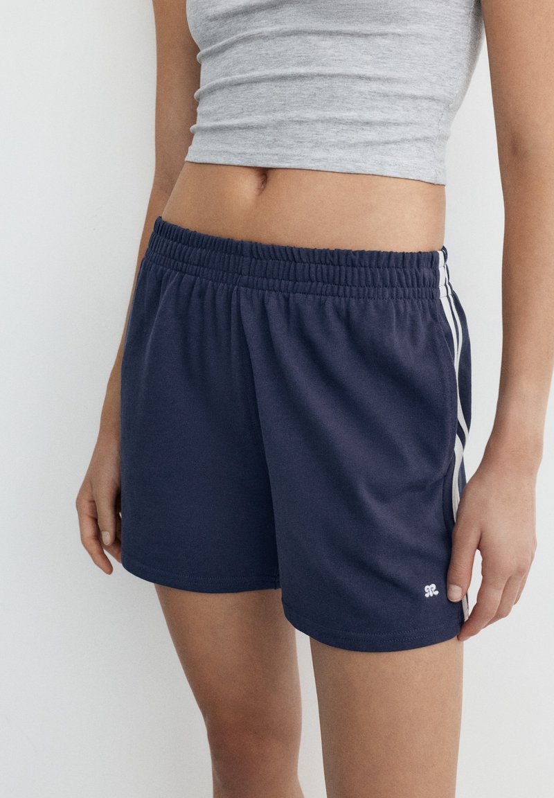 Femme portant un short de sport bleu marine avec des bandes blanches sur les côtés et un débardeur court gris sur un fond blanc uni.