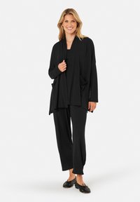 Zwarte lichtgewicht gebreide cardigan met open voorkant en twee voorzakken, gecombineerd met bijpassende zwarte broek, met een relaxte pasvorm en een zachte textuur.