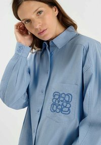 Blauw gestreepte blouse met knoopsluiting en een borstzak, voorzien van gedurfde blauwe borduursels. Zachte stof met een relaxte pasvorm en lange mouwen.