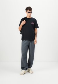 Zwarte katoenen T-shirt met een kleurrijk ontwerp op de borst, gecombineerd met losse grijze joggingsbroeken en beige sneakers. Model draagt een zonnebril.