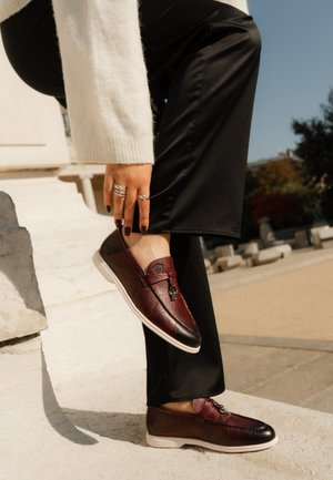 Bordeauxfarbene Leder Slipper mit strukturiertem Finish, schwarzem Zehenkappe und dekorativer Metallbeschläge, kombiniert mit schwarzen Hosen vor einem steinernen Hintergrund.