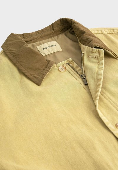 Giacca beige con colletto marrone scuro, cerniera parzialmente aperta, bottone vicino al colletto e etichetta bianca con la scritta "BOBO CHOSES" all'interno della scollatura.