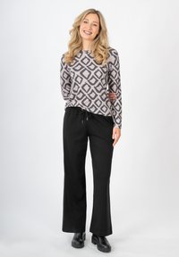 Grauer Langarmoberteil mit einem schwarzen geometrischen Muster, kombiniert mit schwarzen weit geschnittenen Hosen. Das Model trägt schwarze Stiefeletten.