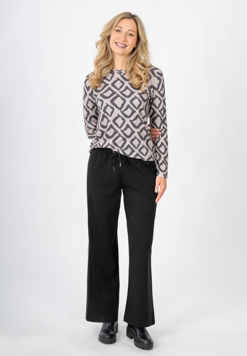 Grauer Langarmoberteil mit einem schwarzen geometrischen Muster, kombiniert mit schwarzen weit geschnittenen Hosen. Das Model trägt schwarze Stiefeletten.