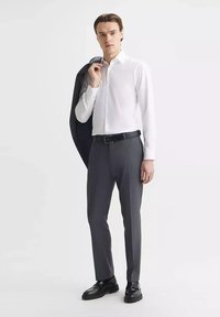 Biała koszula zapinana na guziki, szare spodnie w prążki, czarny pasek i buty. Ciemna kurtka jest narzucona na jedno ramie. Gładki materiał, dopasowany krój.