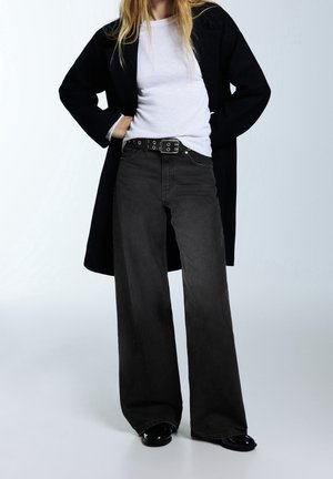 Person wearing a white shirt, black wide-leg jeans with a black belt, black coat, and black shoes, standing with hands on hips.