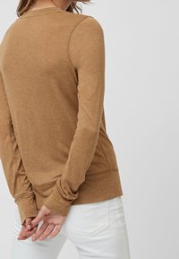 Top marrone a maniche lunghe con un fit rilassato, caratterizzato da polsini a coste e un colletto rotondo, indossato con jeans bianchi. Tessuto morbido e con una texture.