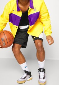 Persona que lleva una chaqueta amarilla y morada de los Lakers, pantalones cortos de baloncesto en blanco y negro, calcetas blancas de Nike y zapatillas de baloncesto en blanco y negro, sosteniendo un balón de baloncesto.