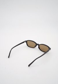 Μαύρα ορθογώνια γυαλιά ηλίου με καφέ φακούς, ανοιχτά επάνω σε λευκή επιφάνεια, το όνομα της μάρκας ορατό στο πλευρικό μπράτσο.