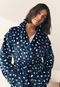 Tamsiai mėlynas vilnos vonios chalatas su baltais taškeliais, ilgomis rankovėmis, šalikiniu apykakliu, surišama diržo juosta ir kišenėmis. Minkšta tekstūra, jaukus dizainas.
