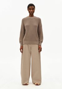 Brauner gestrickter Pullover mit langen Ärmeln, kombiniert mit beigen weit geschnittenen Hosen und lässigen Schuhen mit einem zweifarbigen gelb-roten Design.