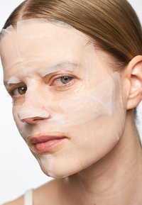 Fuktighetsgivende ansiktsmaske laget av gjennomsiktig materiale, som dekker panne, kinn og haken; huden synlig under med lett naturlig tekstur.