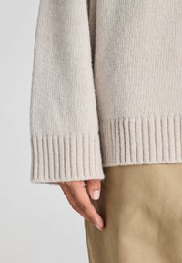 Close-up van een hand naast de zoom en manchetten van een beige gebreide trui, met een achtergrond van beige broek.