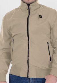 Giacca bomber beige con colletto alto, zip nere, due tasche laterali e un fronte testurizzato. Presenta un'etichetta con logo sul petto sinistro.
