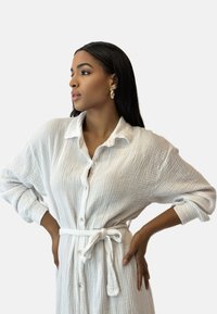 Vestido blanco de botones con tejido_Texturado, que presenta mangas largas y una cintura ceñida con cinturón. Los detalles notables incluyen un cuello y botones plateados.