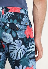 Blomsterprintede bukser i mørkeblå stof med livlige pink og grønne tropiske motiver. Inkluderer en baglomme med knapdetalje.