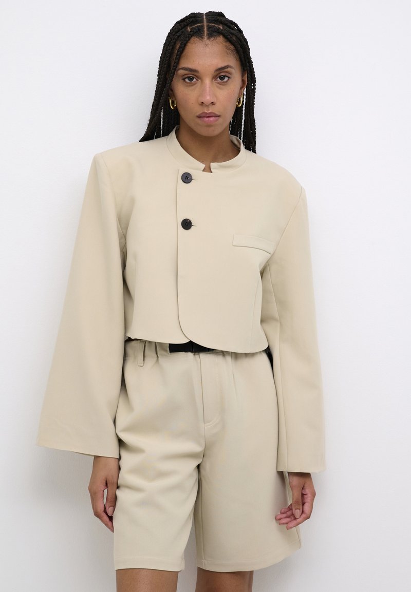 Beige verkürzte Jacke mit rundem Ausschnitt, zwei schwarzen Knöpfen und einer Brusttasche, kombiniert mit passender Shorts und einem schwarzen Gürtel.