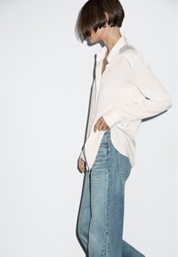 White long-sleeve shirt with a rounded hem, paired with light blue wide-leg denim jeans. Simple design, minimalistic aesthetic.