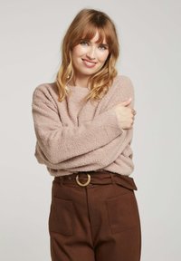 Pull rose clair et duveteux avec de longues manches, porté sur un pantalon marron taille haute avec une large ceinture et une grande boucle. Matière texturée.