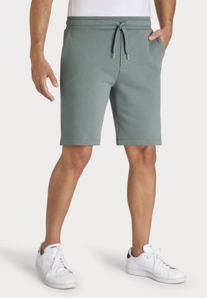 Man wearing grey drawstring shorts with one hand in pocket and white Adidas sneakers on a white background.