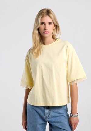 Jonge vrouw met lang blond haar, die een lichtgeel topje met korte mouwen en kanten rand draagt en een blauwe spijkerbroek, staand tegen een effen achtergrond.