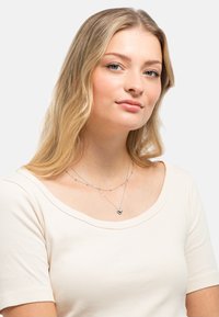 Silberne, mehrlagige Halskette mit einem Herzanhänger, ergänzt durch kleine runde Perlen, getragen über einem hellen, gerippten, cremefarbenen Oberteil.