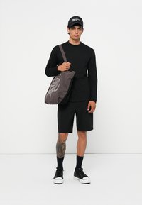 Brun kanvas skulderveske med et stort grafisk design, brukt over en svart langermet skjorte og svarte shorts. Sorte sneakers og caps fullfører looken.