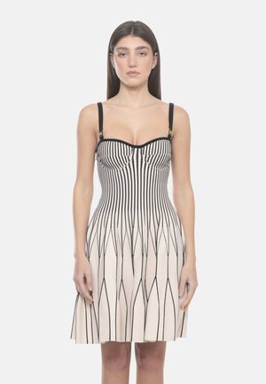 Donna con lunghi capelli castani che indossa un vestito beige senza maniche con righe nere verticali e a zigzag, in piedi davanti a uno sfondo uniforme.