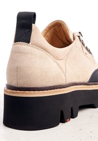 Scarpa in suede beige con tacco e suola neri a contrasto. Presenta una finitura testurizzata, lacci resistenti e un accento rosso visibile sulla suola.