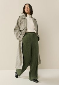Checkered trench coat in beige, blue, and green; cream turtleneck; wide-legged green corduroy pants; black shoes; smooth textures.