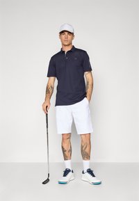 Námornícky polo tričko s golierom, biele šortky a biele golfové topánky s námorníckymi akcentmi. Drží golfovú palicu. Tetovania sú viditeľné na ramenách.