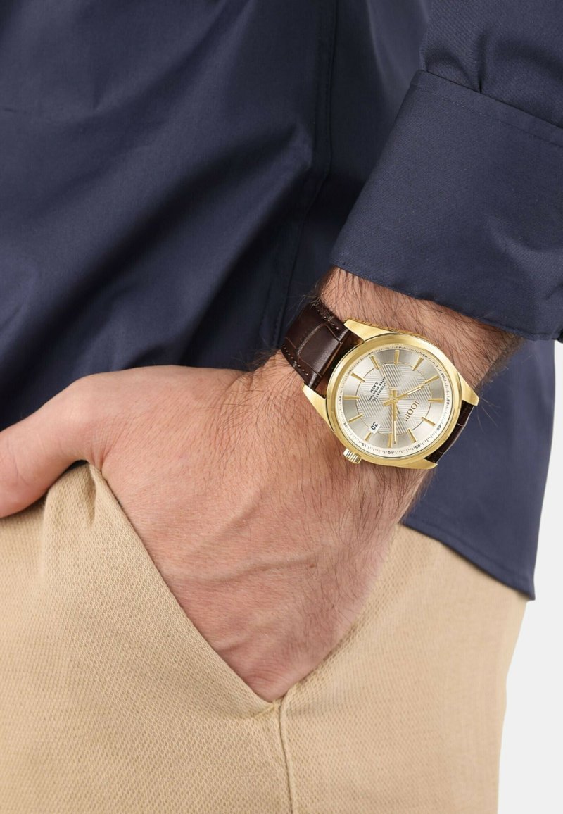 Mans Handgelenk, das eine goldene Uhr mit einem braunen Lederarmband trägt, Hand in der Tasche einer beigen Hose, langärmliges navyblaues Hemd sichtbar.